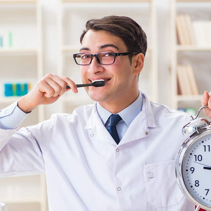 Man in a white coat brushing his teeth and holding an alarm clock, with a blurred indoor background.