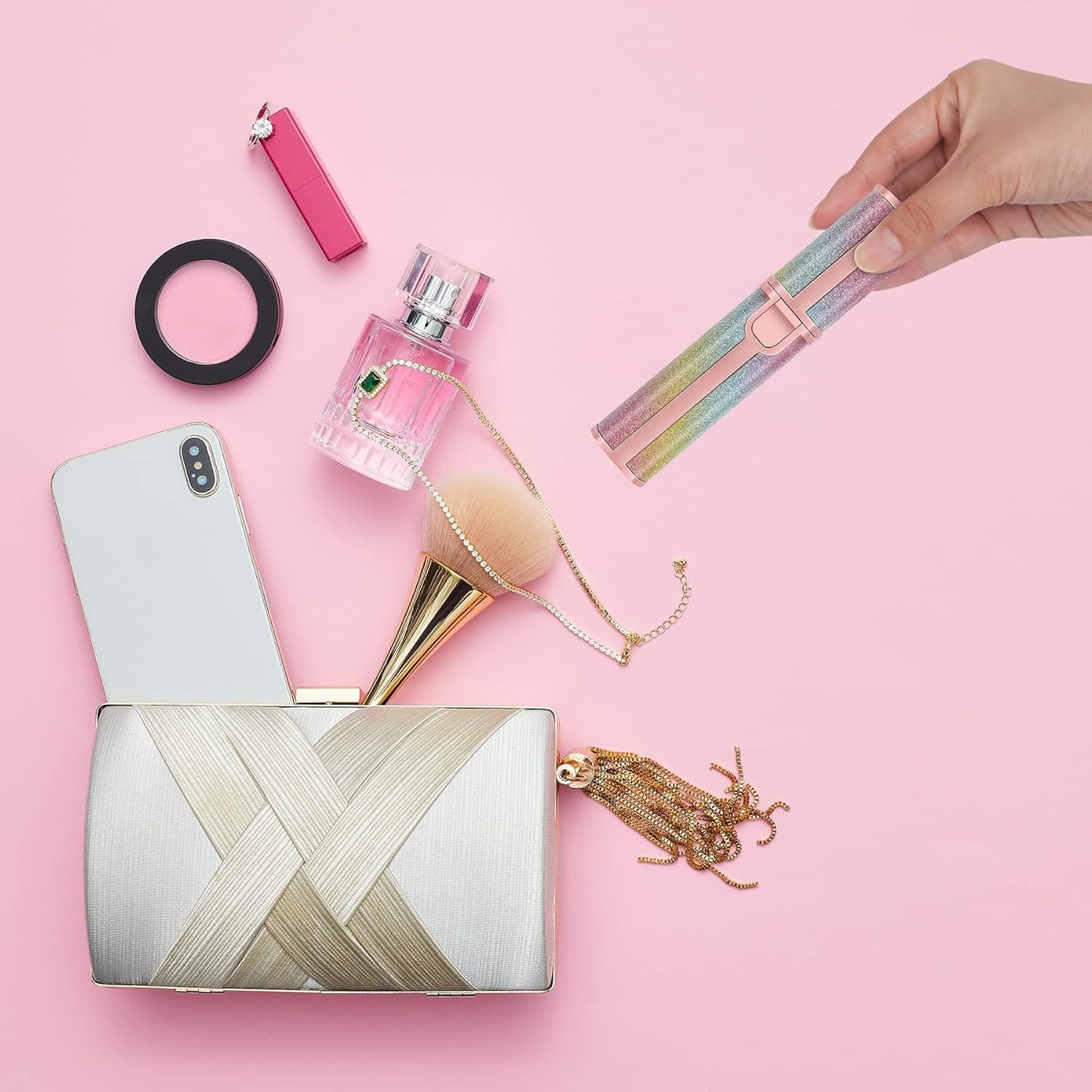 Collection of beauty products including a phone, perfume, brush, and clutch on a pink background