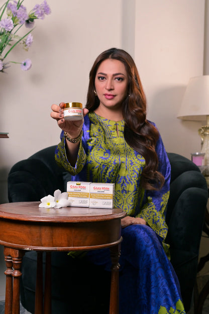 Woman holding a skincare product in a well-lit room with a table and flowers in the background