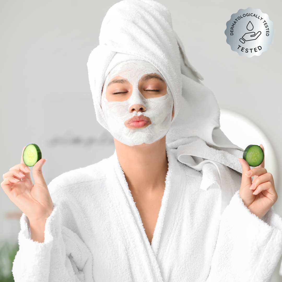 Person with a facial mask holding cucumber slices, wearing a towel and robe, with a dermatologically tested logo.
