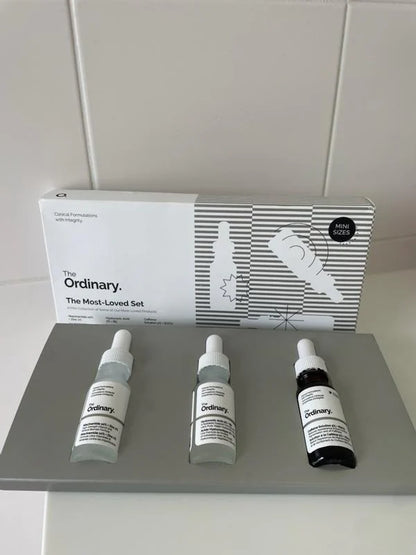The Ordinary The Most-Loved Set displayed with its retail box and three mini skincare serums arranged on a gray platform. The set includes Niacinamide 10% + Zinc 1%, Hyaluronic Acid 2% + B5, and Caffeine Solution 5% + EGCG, designed for hydration, blemish care, and under-eye support.