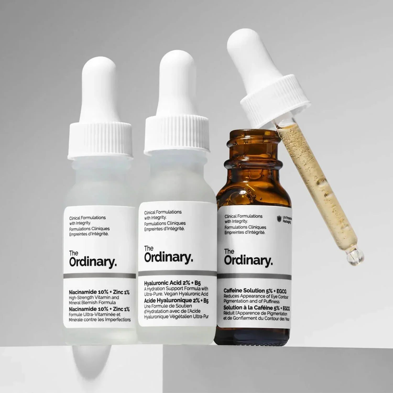 Three mini-sized The Ordinary skincare serums displayed together, including Niacinamide 10% + Zinc 1%, Hyaluronic Acid 2% + B5, and Caffeine Solution 5% + EGCG. An open amber bottle with a serum-filled dropper highlights the collection’s hydration, blemish-care, and eye-care benefits.